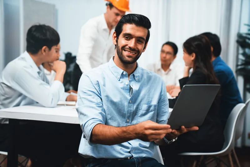 Jefe de capacitación de una empresa muestra en su laptop el aula virtual al equipo, con un supervisor de seguridad con casco al fondo