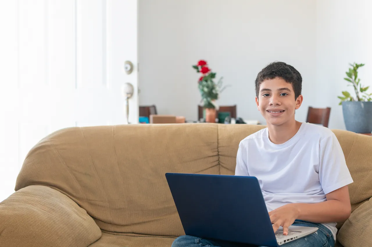 Alumno de secundaria estudiando en casa con su laptop en el sofá, conectado a su colegio virtual