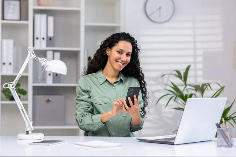 Asesora de admisión de un instituto respondiendo a interesados por WhatsApp desde su celular, con la laptop del CRM sobre el escritorio