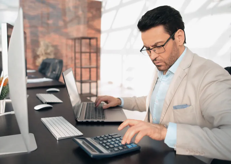 Director de un centro de capacitación calculando en su escritorio cuánto cobrar por un curso online, con calculadora y laptop