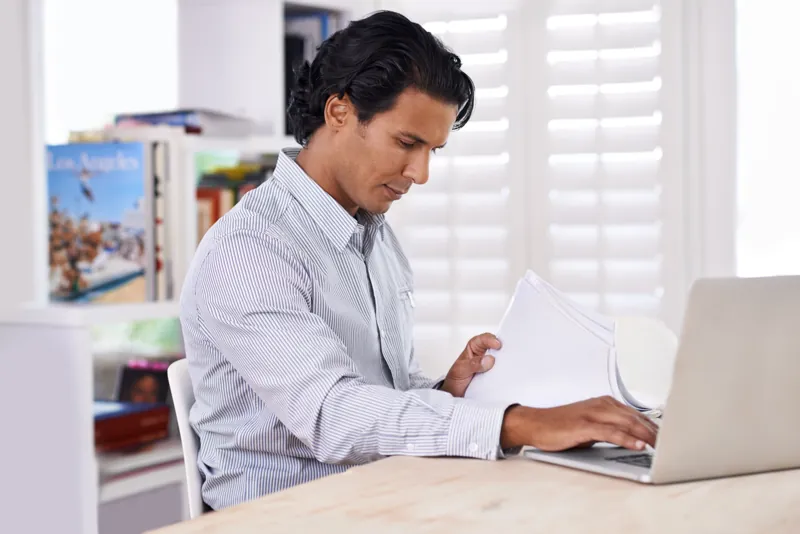Dueño de una pequeña empresa revisando en su laptop y en papel los costos anuales de mantener su página web