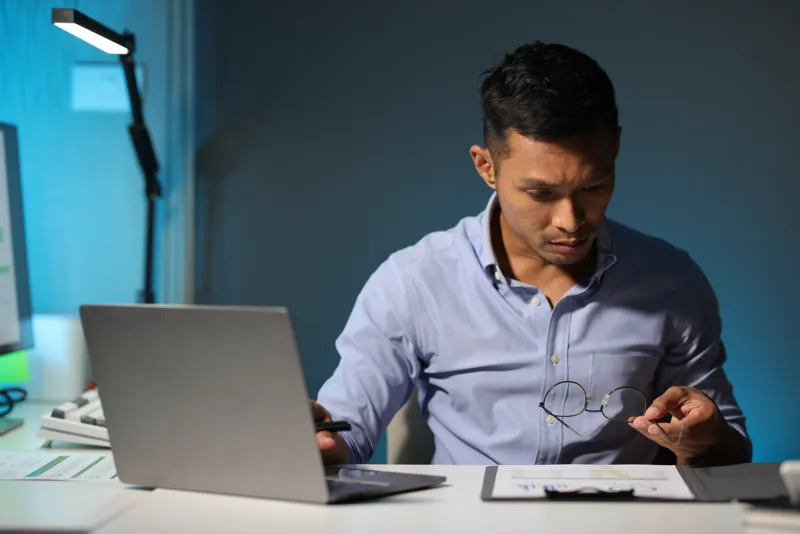 Dueño de una empresa revisando en su laptop el presupuesto de un sistema a medida, con documentos y lentes en la mano