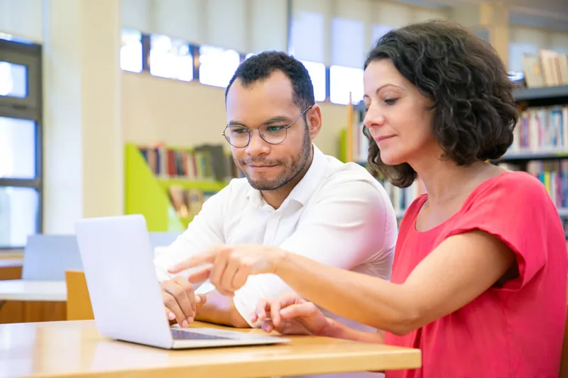 Docente de un instituto revisando con un alumno adulto un trabajo en la laptop, en una biblioteca, tema inteligencia artificial en la educación