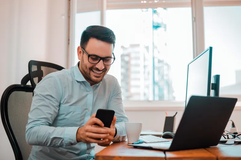 Empresario sonriente con lentes revisando en su celular y su laptop las ventas de cursos cobradas con Mercado Pago