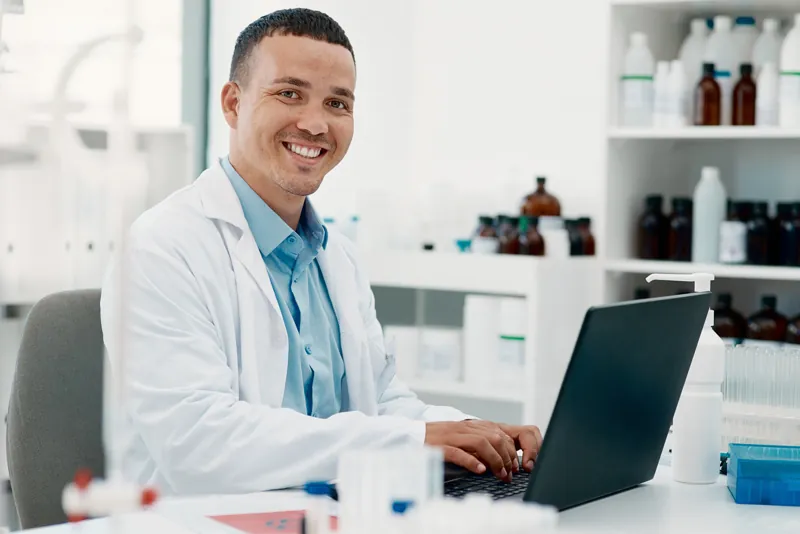 Tecnólogo médico sonriendo en un laboratorio clínico mientras registra resultados en el sistema desde su laptop