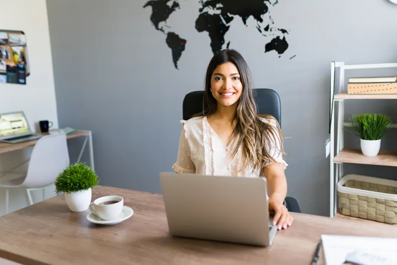 Asesora de una agencia de viajes sonriendo en su escritorio frente a la laptop con el sistema de reservas online, con un mapa del mundo detrás