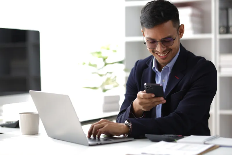 Director de una empresa respondiendo mensajes de WhatsApp Business en su celular frente a la laptop en su oficina