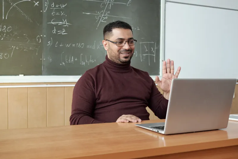 Docente saludando a sus alumnos frente a la laptop durante una clase en vivo, con una pizarra con fórmulas detrás